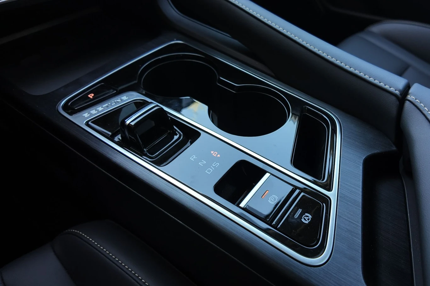 Nowoczesna konsola środkowa samochodu z elektroniczną dźwignią zmiany biegów, dwoma uchwytami na kubki i przyciskami funkcyjnymi, otoczona czarnym, błyszczącym wykończeniem i jasnymi przeszyciami na tapicerce.