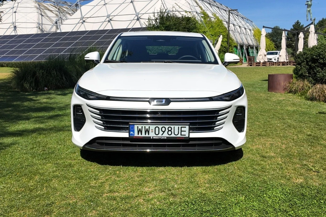 Biały samochód elektryczny stoi na zielonym trawniku, na tle nowoczesnych budynków i paneli fotowoltaicznych, w słoneczny dzień.