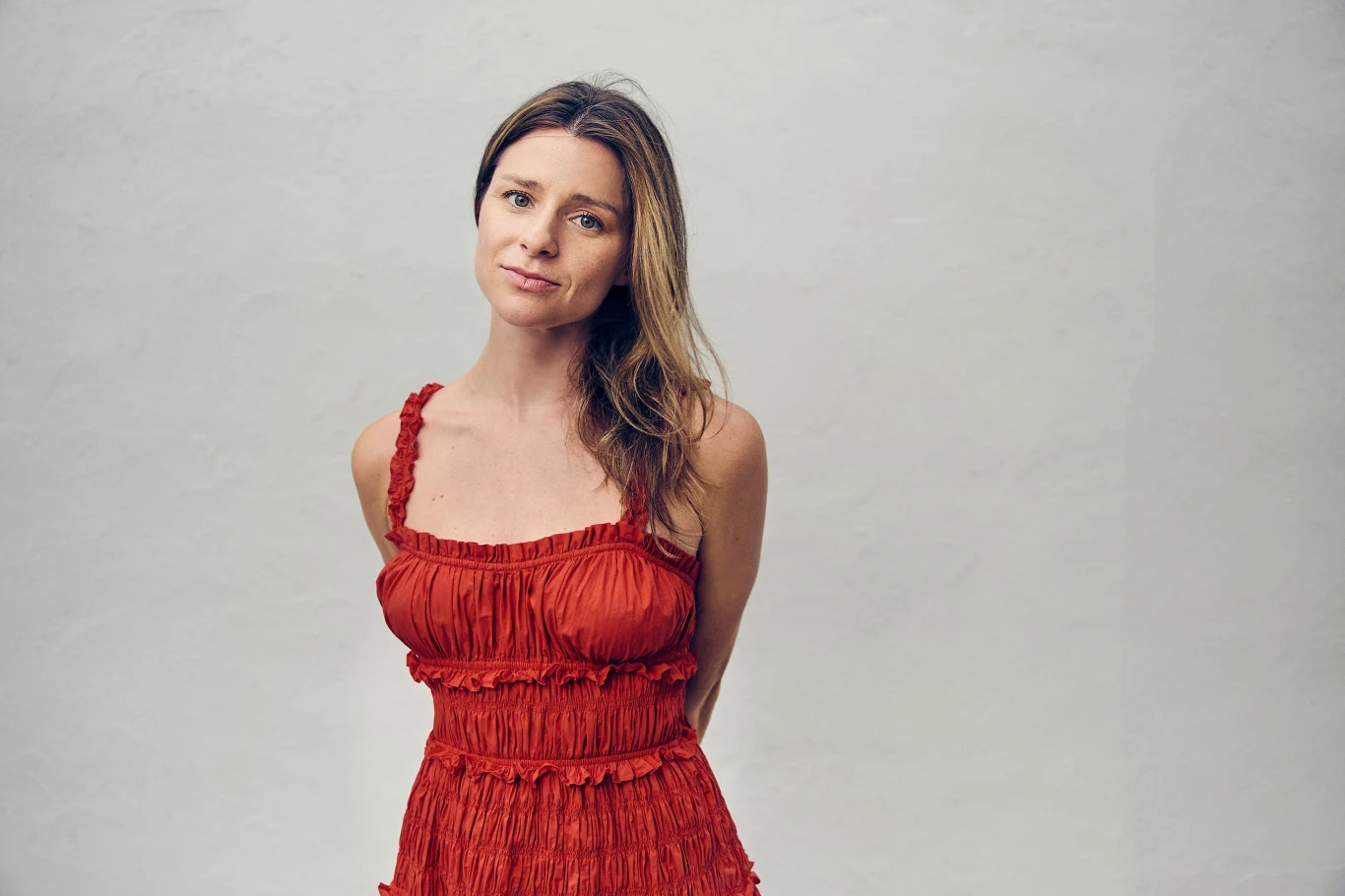 Lee Tilghman Kobieta w czerwonej sukience patrząca spokojnie w stronę aparatu, stojąca na jasnym, jednolitym tle, z rękami schowanymi za plecami i delikatnym uśmiechem.