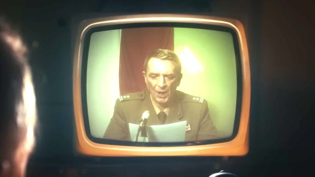 Cronos: The New Dawn - screen z trailera gry Wojskowy w mundurze siedzi przed mikrofonem i odczytuje oświadczenie, sylwetka mężczyzny wyświetlona na ekranie starego telewizora, w tle widoczna dwukolorowa flaga.