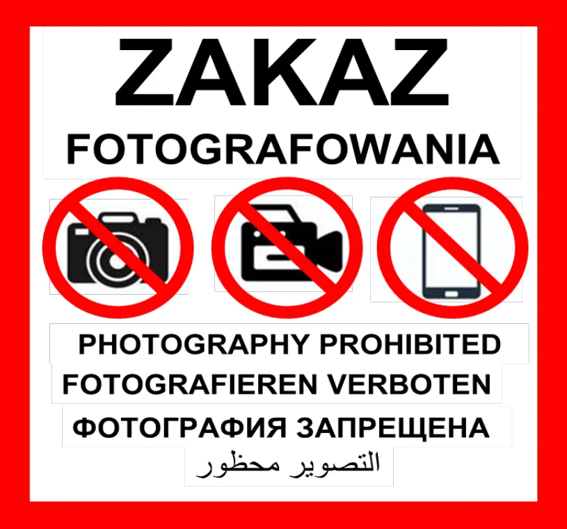 Zakaz fotografowania wyrażony za pomocą symboli przekreślonych aparatu, kamery i telefonu, otoczonych czerwonym obramowaniem oraz napisu w kilku językach, w tym po polsku, angielsku, niemiecku, rosyjsku i arabsku.