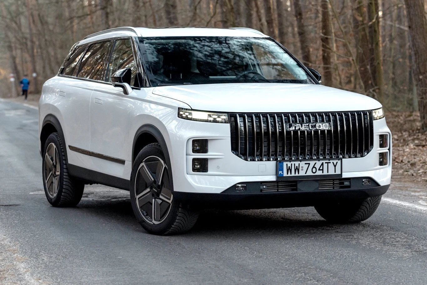 Jaecoo 7 Biały nowoczesny SUV marki Leapmotor z charakterystycznym dużym chromowanym grillem, poruszający się po asfaltowej drodze w lesie, w tle widoczne drzewa i osoba biegnąca po lewej stronie jezdni.