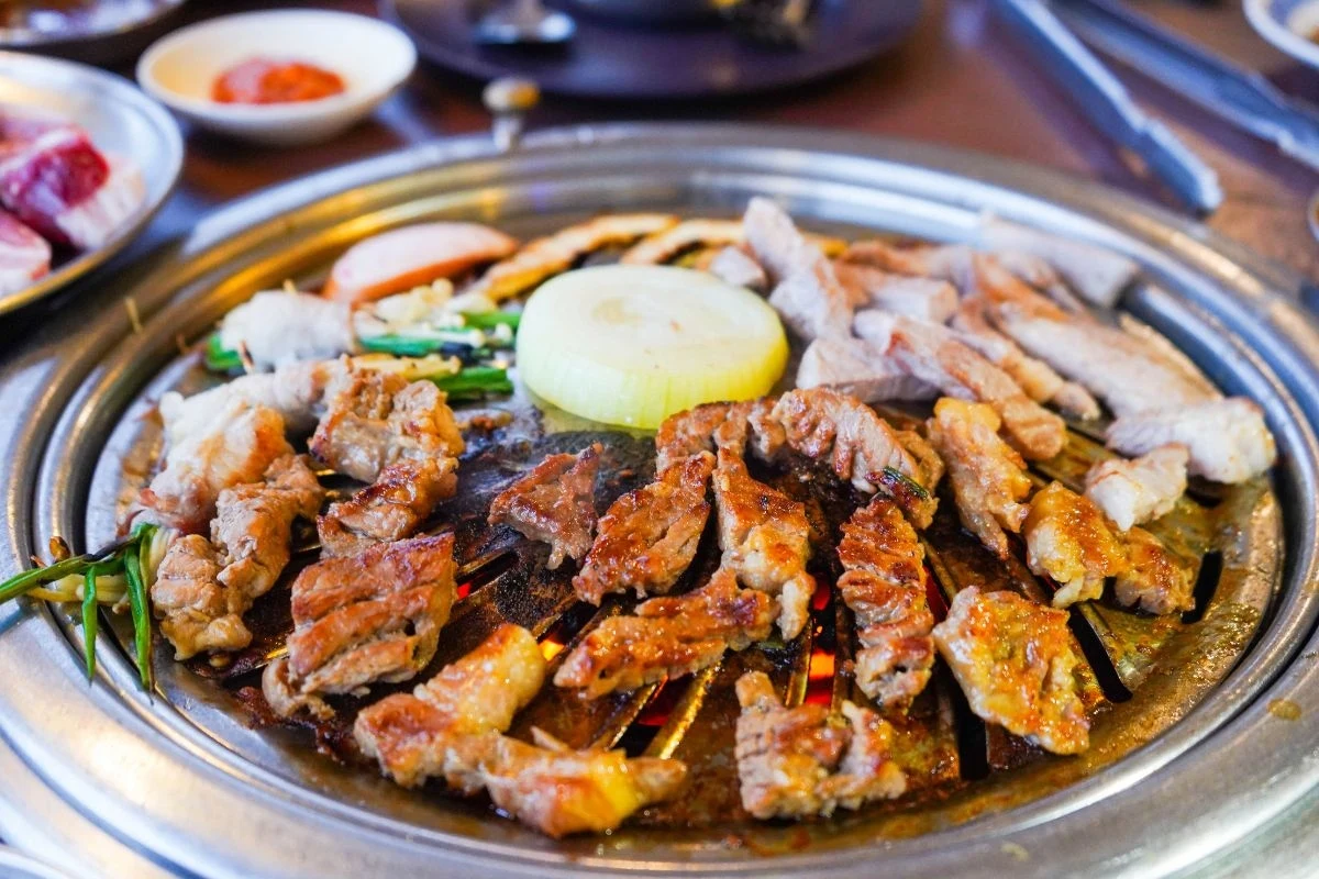 Sezon grillowy dobiega końca. Jak pomóc wątrobie w regeneracji? Różne kawałki mięsa grillowane razem z warzywami takimi jak cebula oraz zielone pędy, na okrągłej płycie grillowej, podane z dodatkami w tle.
