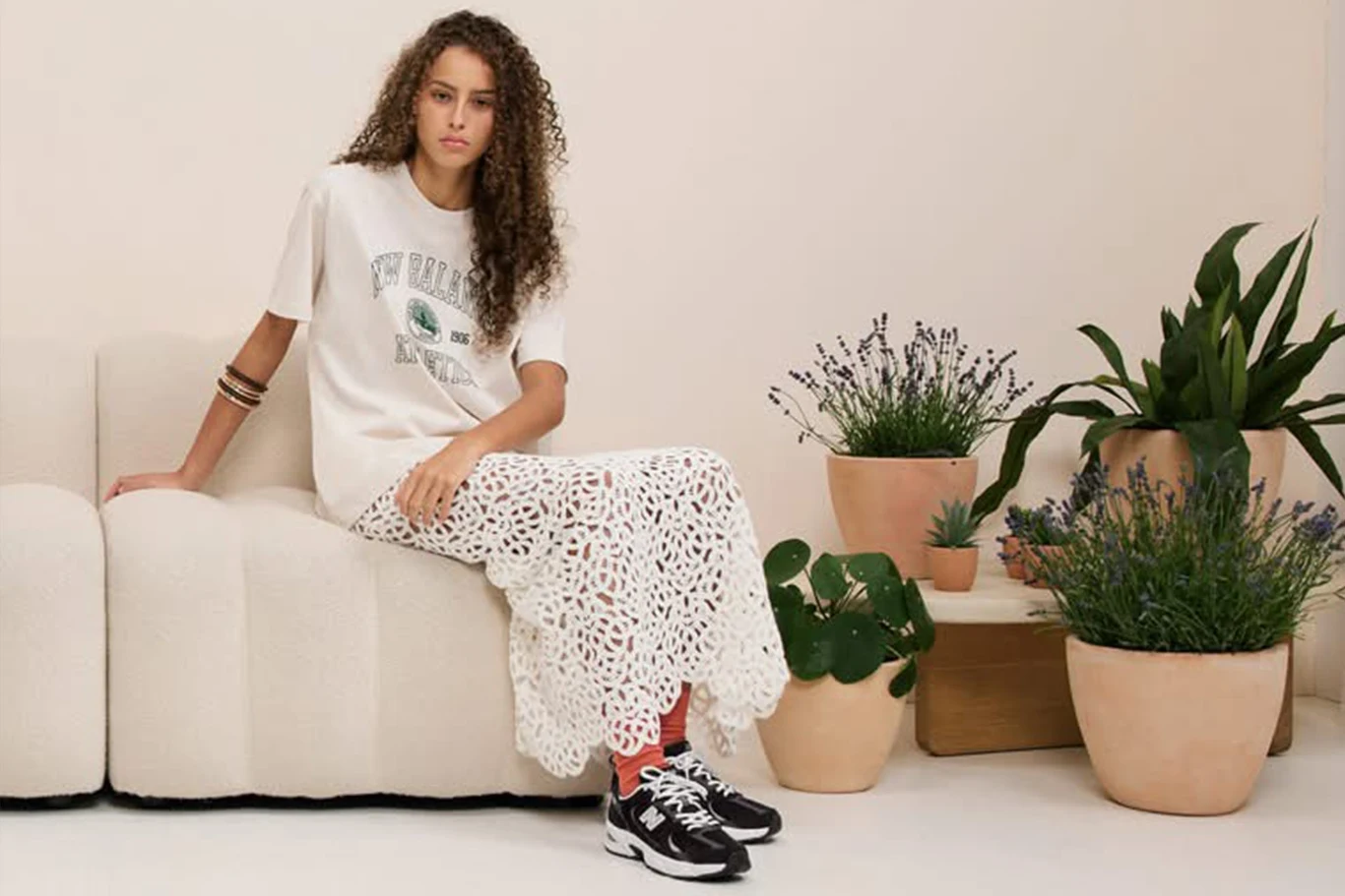 Młoda kobieta siedzi na jasnej kanapie, ubrana w t-shirt i koronkową spódnicę, z czarnymi butami New Balance na nogach.