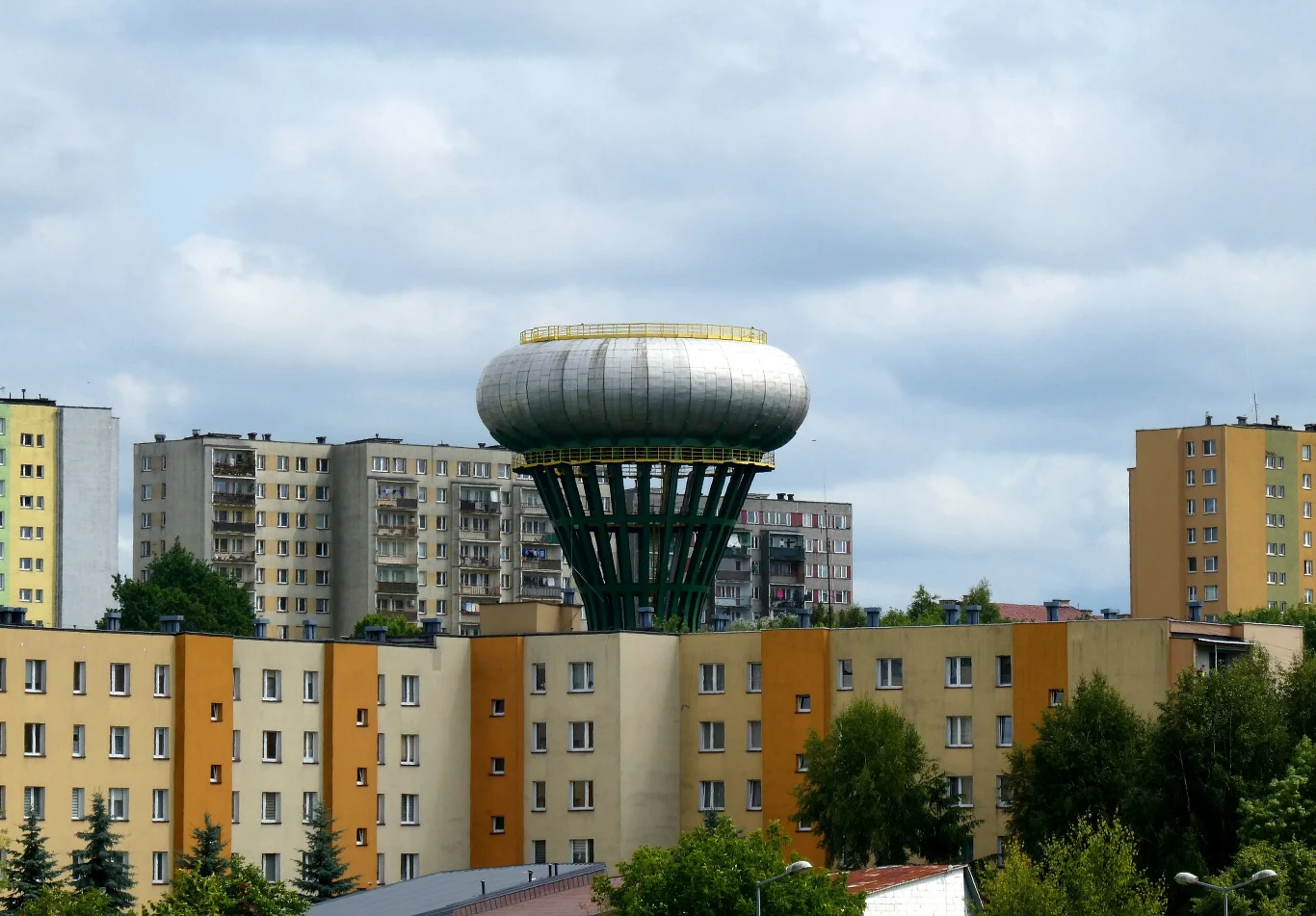Charakterystyczny okrągły zbiornik wodny osadzony na betonowej konstrukcji, otoczony blokami mieszkalnymi z wielkiej płyty oraz drzewami, pod lekko zachmurzonym niebem.