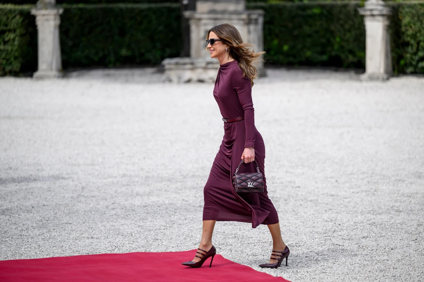 Kobieta ubrana w elegancką bordową sukienkę i wysokie szpilki idzie po czerwonym dywanie, trzymając w dłoni stylową torebkę; w tle widoczne zadbane krzewy oraz elementy architektury ogrodowej.