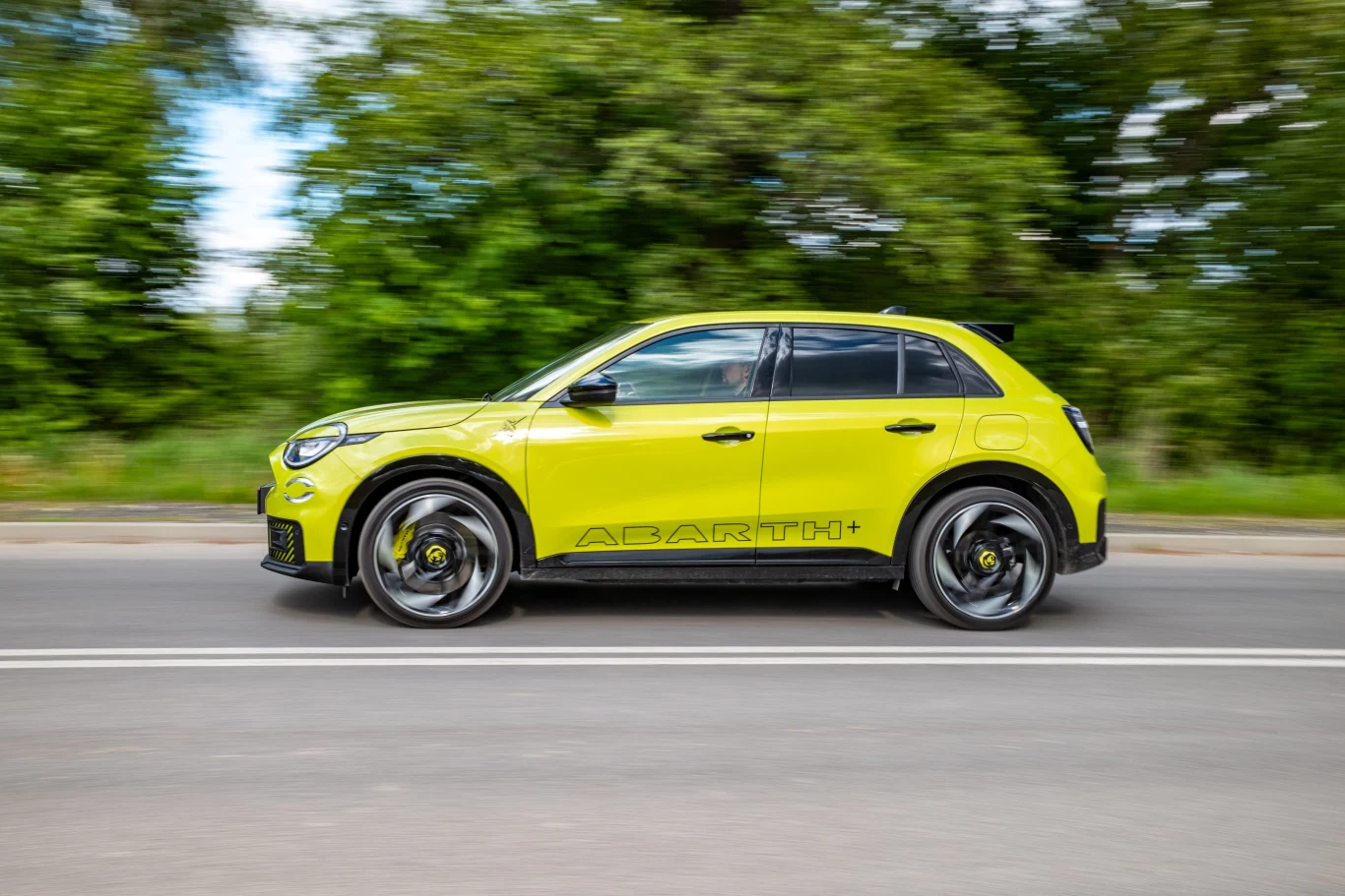 Jaskrawożółty samochód typu SUV marki Abarth porusza się po drodze asfaltowej, w tle zielone drzewa.