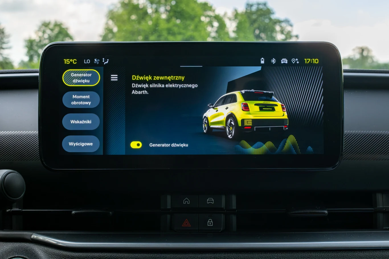 Wyświetlacz samochodowy z widoczną grafiką żółtego samochodu elektrycznego Abarth oraz menu po lewej stronie, z przyciskami dotyczącymi generowania dźwięku i parametrów jazdy, w tle drzewa i niebo widoczne przez przednią szybę.