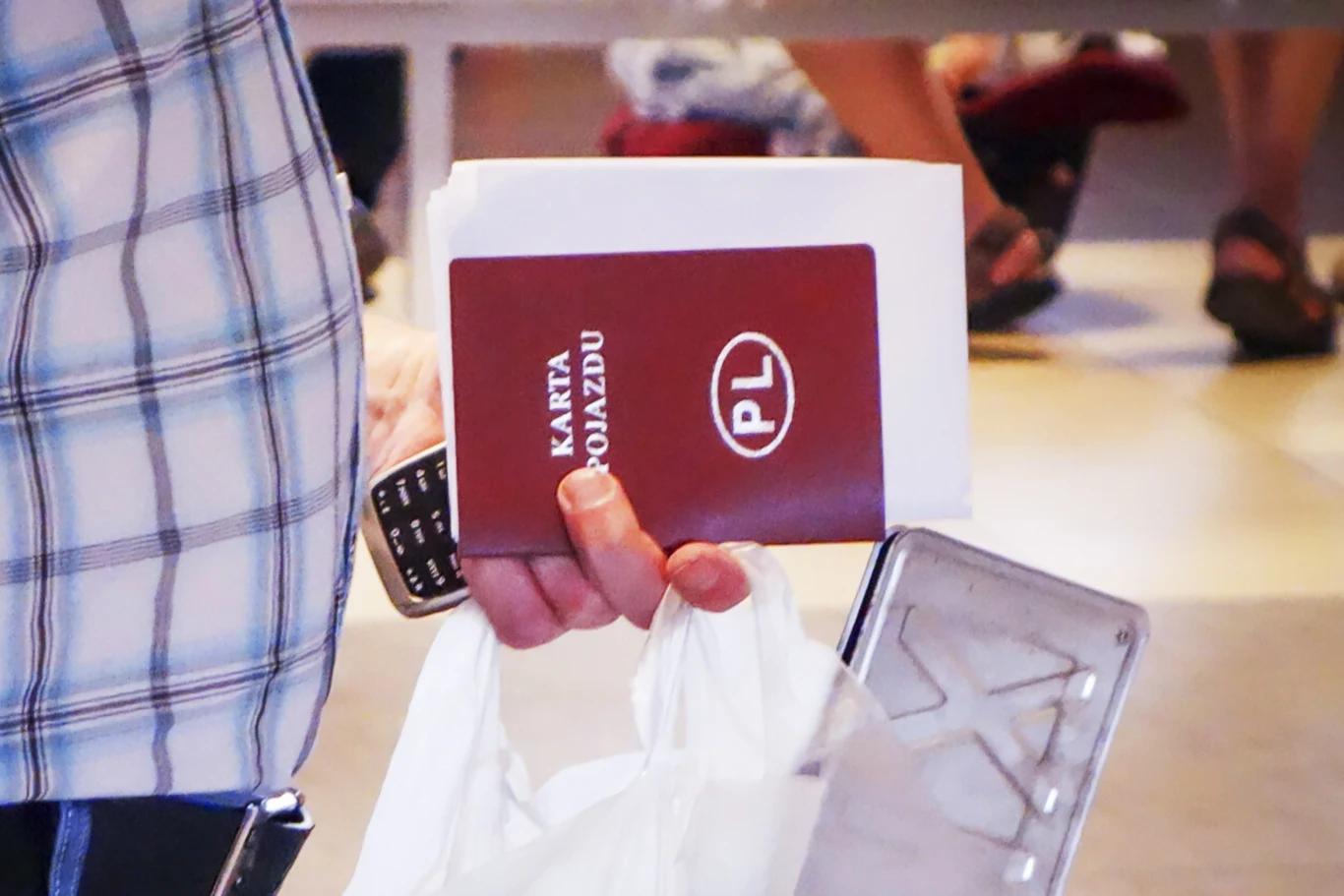 Dłoń trzymająca polski dokument samochodowy 'Karta Pojazdu', plastikową torbę z zawartością oraz obiekt przypominający tablicę rejestracyjną, w tle rozmyte postacie i podłoga.