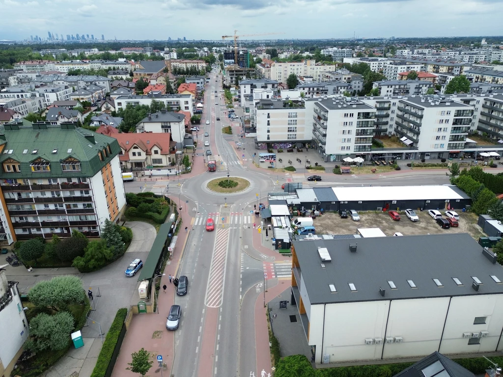 Skrzyżowanie dróg z rondem otoczone budynkami mieszkalnymi i usługowymi, samochody poruszające się po jezdni, wyraźnie widoczne miejsca parkingowe, szerokie ścieżki rowerowe, estetyczna i nowoczesna zabudowa miejska z dużą ilością zieleni.
