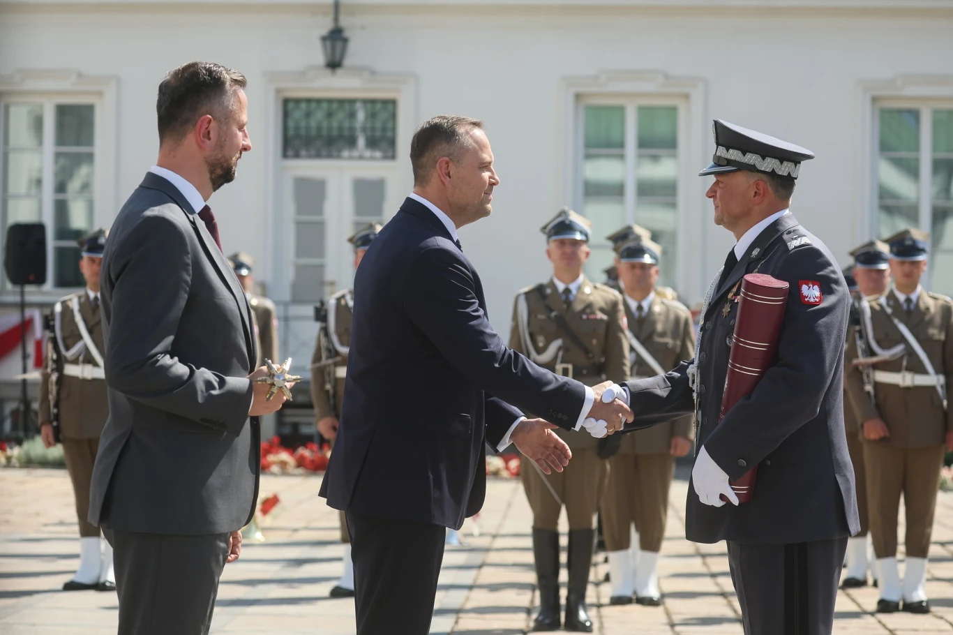 Prezydent Karol Nawrocki wręczył nominacje generalskie Oficjalne spotkanie z udziałem prezydenta, wojskowego i urzędnika, w tle stoi szereg umundurowanych żołnierzy w paradnym szyku, wręczanie dokumentu oraz wymiana uścisku dłoni podczas ceremonii państwowej.