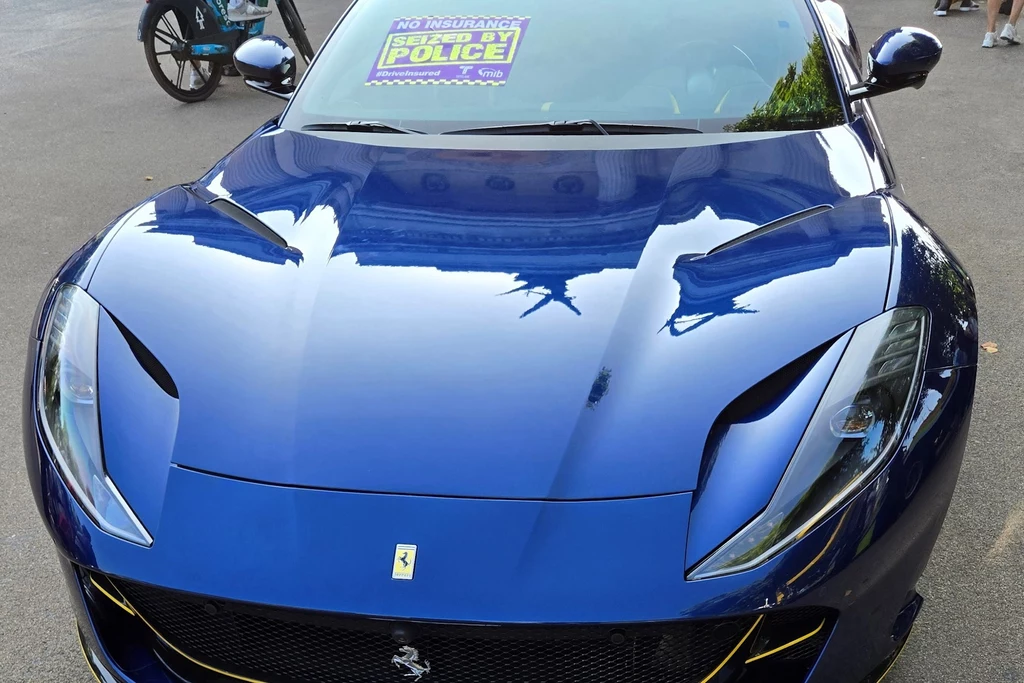 Niebieski sportowy samochód Ferrari zaparkowany na ulicy, na przedniej szybie znajduje się żółta kartka z napisem informującym o braku ubezpieczenia oraz interwencji policji.