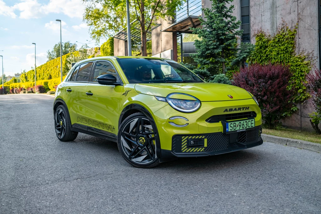 Abarth 600e ma łączyć sportowe wrażenia z jazdy z codzienną praktycznością Jaskrawożółty samochód osobowy marki Abarth zaparkowany na ulicy w zielonej, zadbanej okolicy, otoczony roślinnością i budynkami mieszkalnymi.