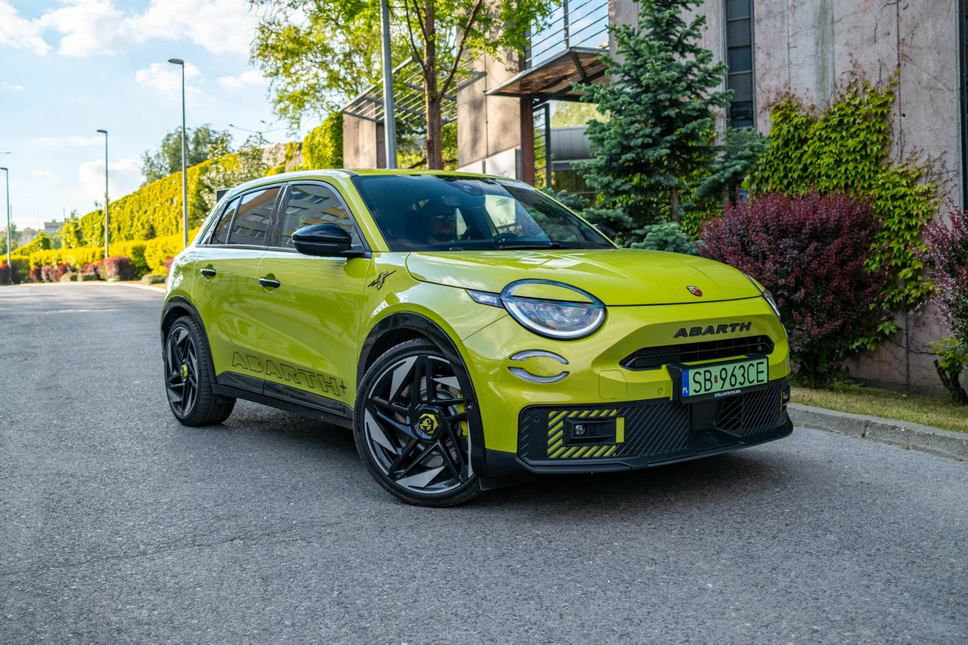 Jaskrawożółty samochód osobowy marki Abarth zaparkowany na ulicy w zielonej, zadbanej okolicy, otoczony roślinnością i budynkami mieszkalnymi.