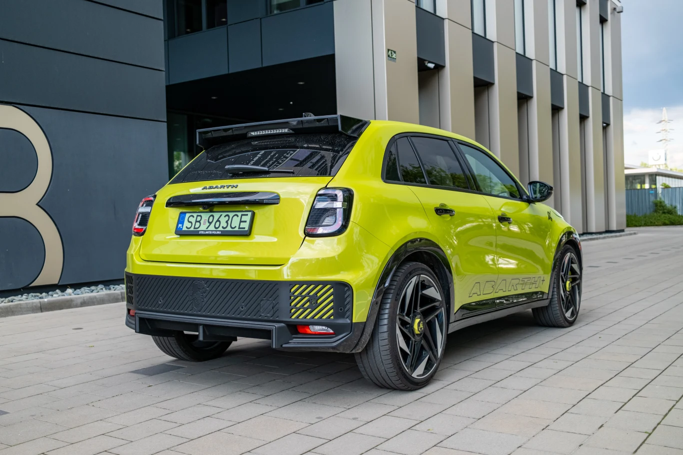 Jasnozielony, sportowy samochód marki Abarth zaparkowany na tle nowoczesnego budynku, widok z tyłu pod lekkim kątem, podkreślający sportowy wygląd i detale nadwozia oraz felgi.