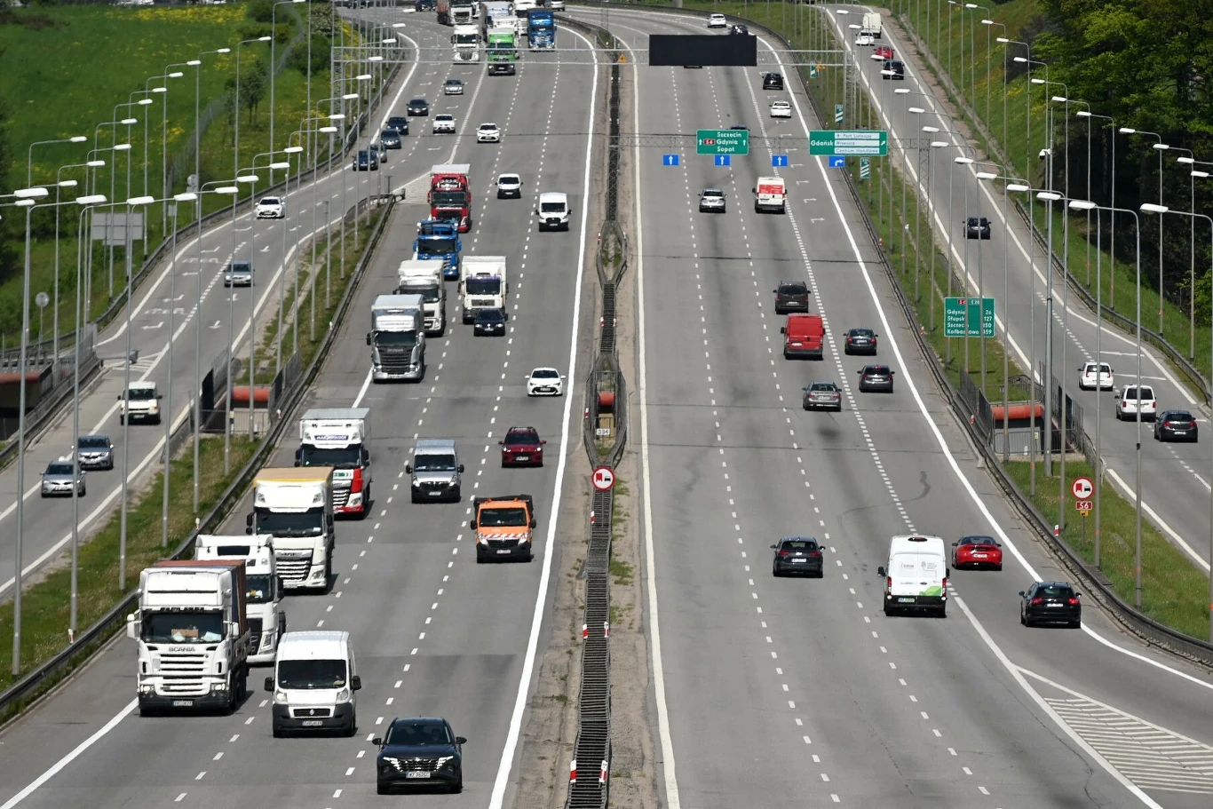 Polskie drogi są coraz lepsze, samochody również coraz bezpieczniejsze Autostrada z wieloma pasami ruchu, po których poruszają się samochody osobowe i ciężarówki, dwie przeciwległe jezdnie oddzielone barierą, po obu stronach latarnie i zielone pobocza.