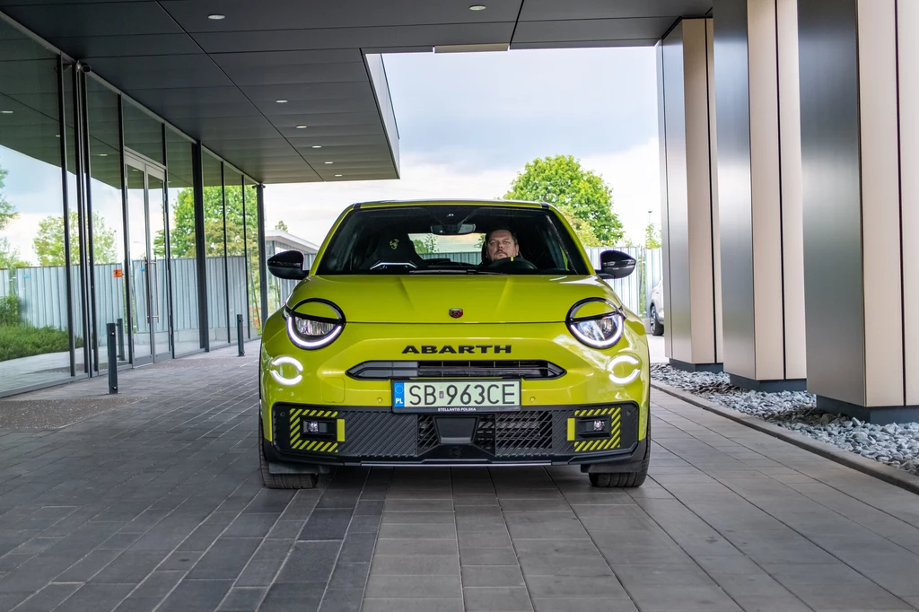 Abarth 600e Scorpionissima Jaskrawy, żółty samochód marki Abarth z charakterystycznym agresywnym designem stoi pod nowoczesnym budynkiem, kierowca widoczny za kierownicą, w tle drzewa i szklana elewacja.