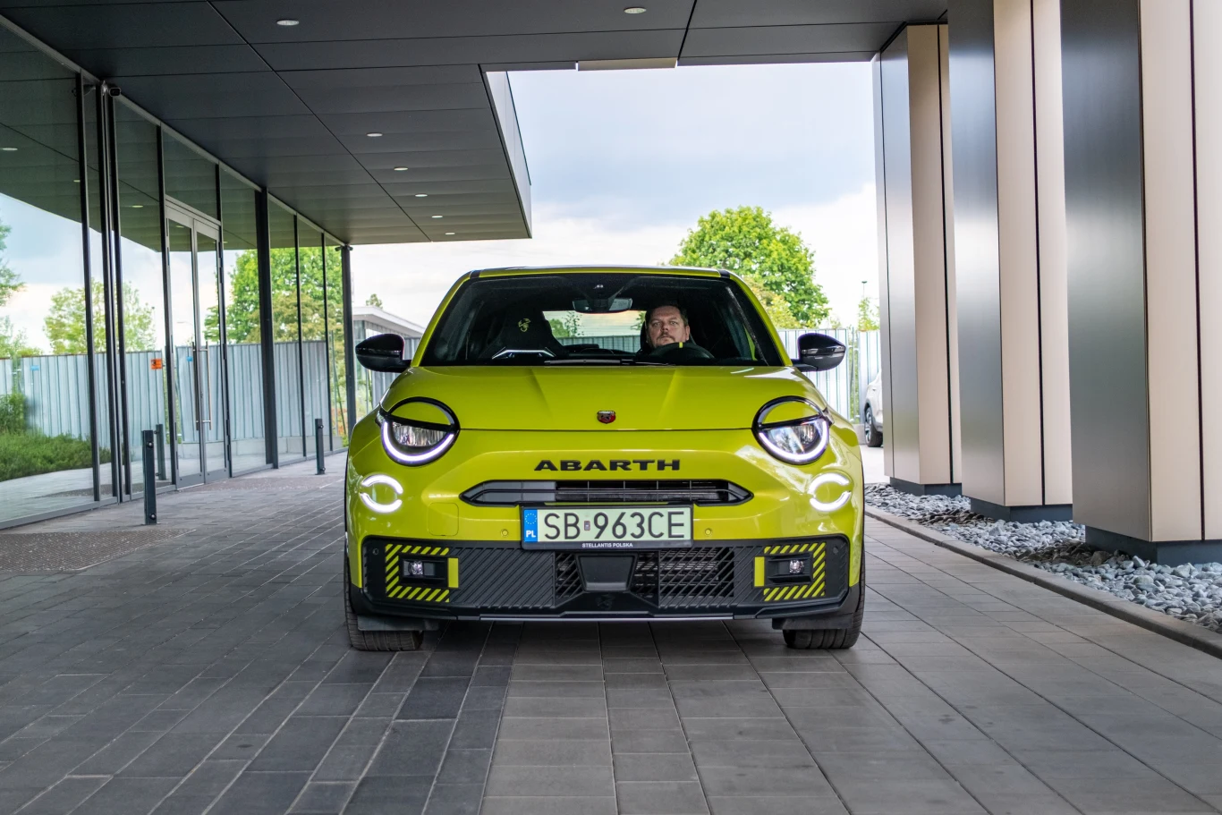 Jaskrawy, żółty samochód marki Abarth z charakterystycznym agresywnym designem stoi pod nowoczesnym budynkiem, kierowca widoczny za kierownicą, w tle drzewa i szklana elewacja.
