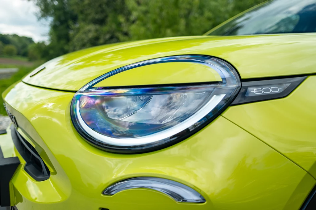 Abarth 600e Scorpionissima Zbliżenie na reflektor przedni nowoczesnego, żółtego samochodu elektrycznego z wyraźnie widocznym oznaczeniem modelu E600, w tle rozmyta zieleń.