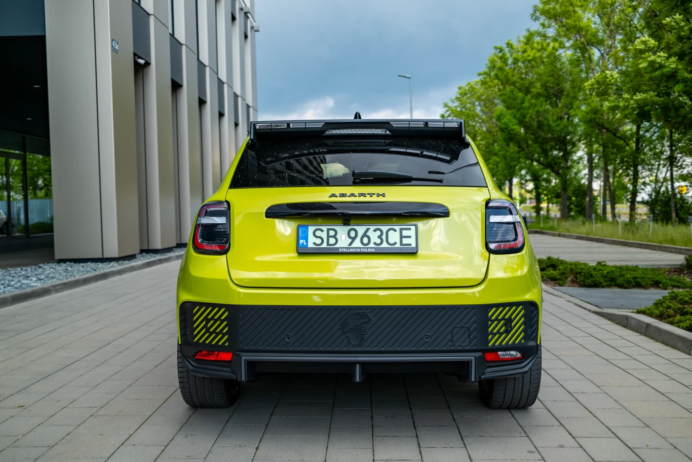 Jaskrawozielony samochód marki Abarth zaparkowany tyłem na brukowanej alejce obok nowoczesnego budynku i zielonych drzew, widoczna tablica rejestracyjna i charakterystyczne sportowe detale auta.