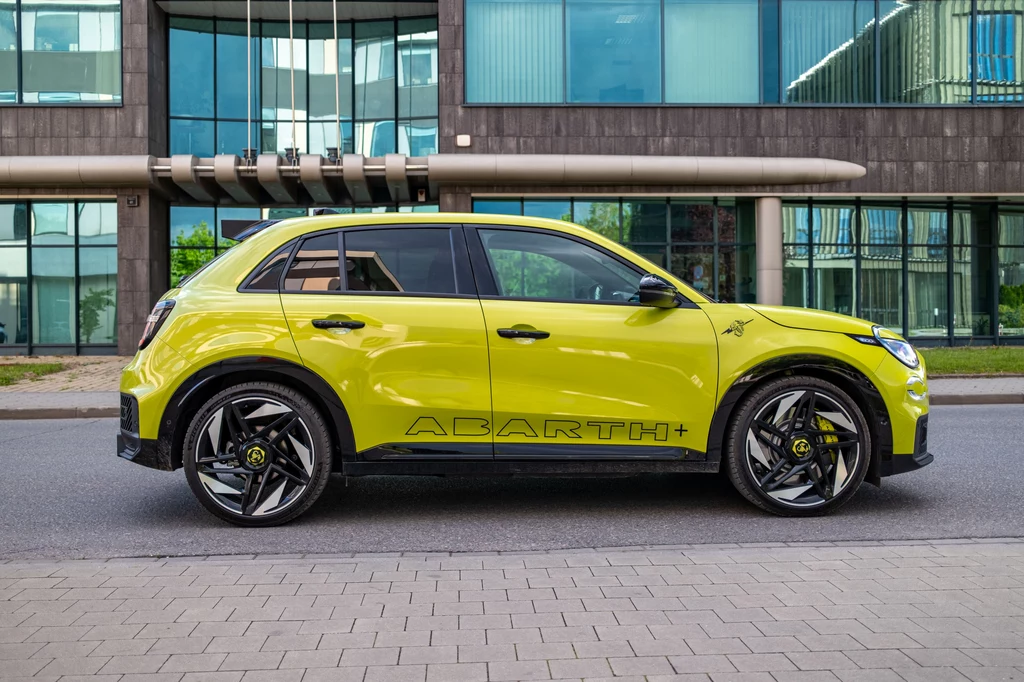 Abarth 600e Scorpionissima Jaskrawożółty nowoczesny samochód terenowy typu SUV marki Abarth zaparkowany przy ulicy na tle nowoczesnego biurowca z dużymi przeszkleniami.
