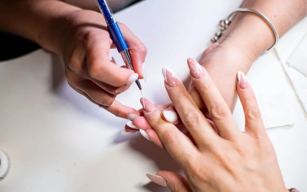 Używasz tych lakierów? 1 września musisz je wyrzucić Ręce osoby wykonującej manicure; jedna ręka trzyma dłoń klientki, druga używa niebieskiego przyrządu do ozdabiania paznokci, paznokcie są długie, pomalowane na biało-różowo, w tle widoczne fragmenty biżuterii i jasne powierzchnie.