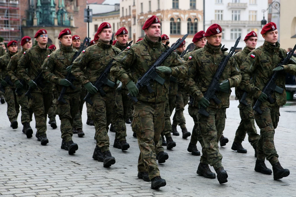 Oddział żołnierzy w mundurach wojskowych i czerwonych beretach maszeruje w zwartej formacji z karabinami w rękach po brukowanej ulicy miasta, w tle widoczne budynki oraz fragmenty historycznej architektury.