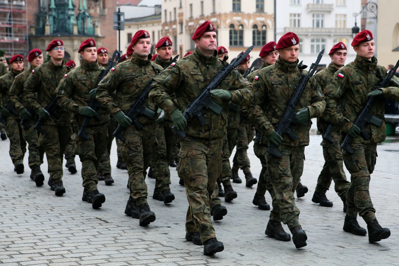 Oddział żołnierzy w mundurach wojskowych i czerwonych beretach maszeruje w zwartej formacji z karabinami w rękach po brukowanej ulicy miasta, w tle widoczne budynki oraz fragmenty historycznej architektury.