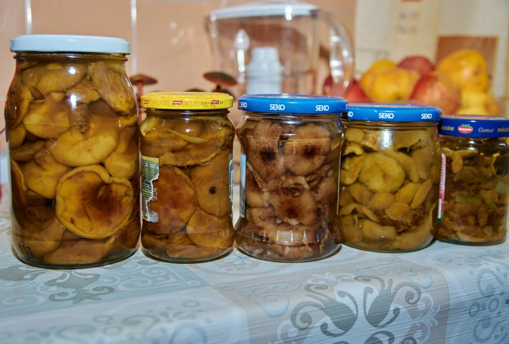 Nie marynuj robaczywych grzybów. Szklane słoiki wypełnione przetworami z plasterków jabłek ustawione w rzędzie na stole, w tle przezroczysty dzbanek i grupka świeżych jabłek.