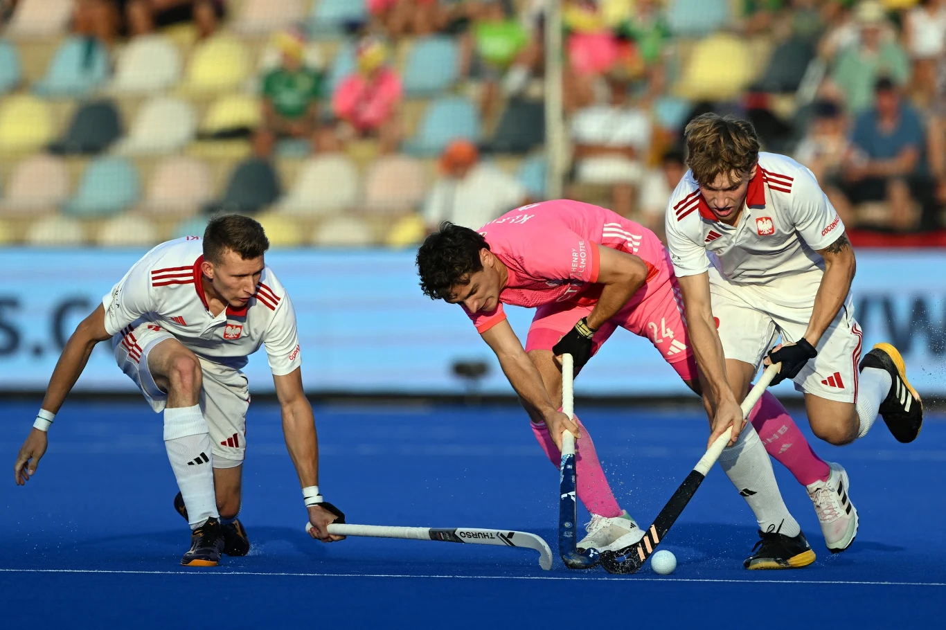 Dwóch zawodników w białych strojach oraz jeden w różowym rywalizują o piłeczkę do hokeja na niebieskim boisku, wszyscy są skoncentrowani na grze, a w tle widoczna jest rozmyta publiczność na trybunach.