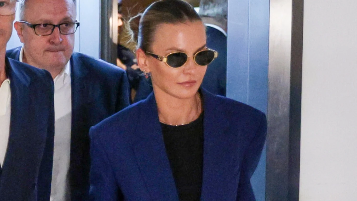 Kobieta w okularach przeciwsłonecznych i granatowej marynarce stoi obok mężczyzny w garniturze, oboje wchodzą przez drzwi, tło rozmyte