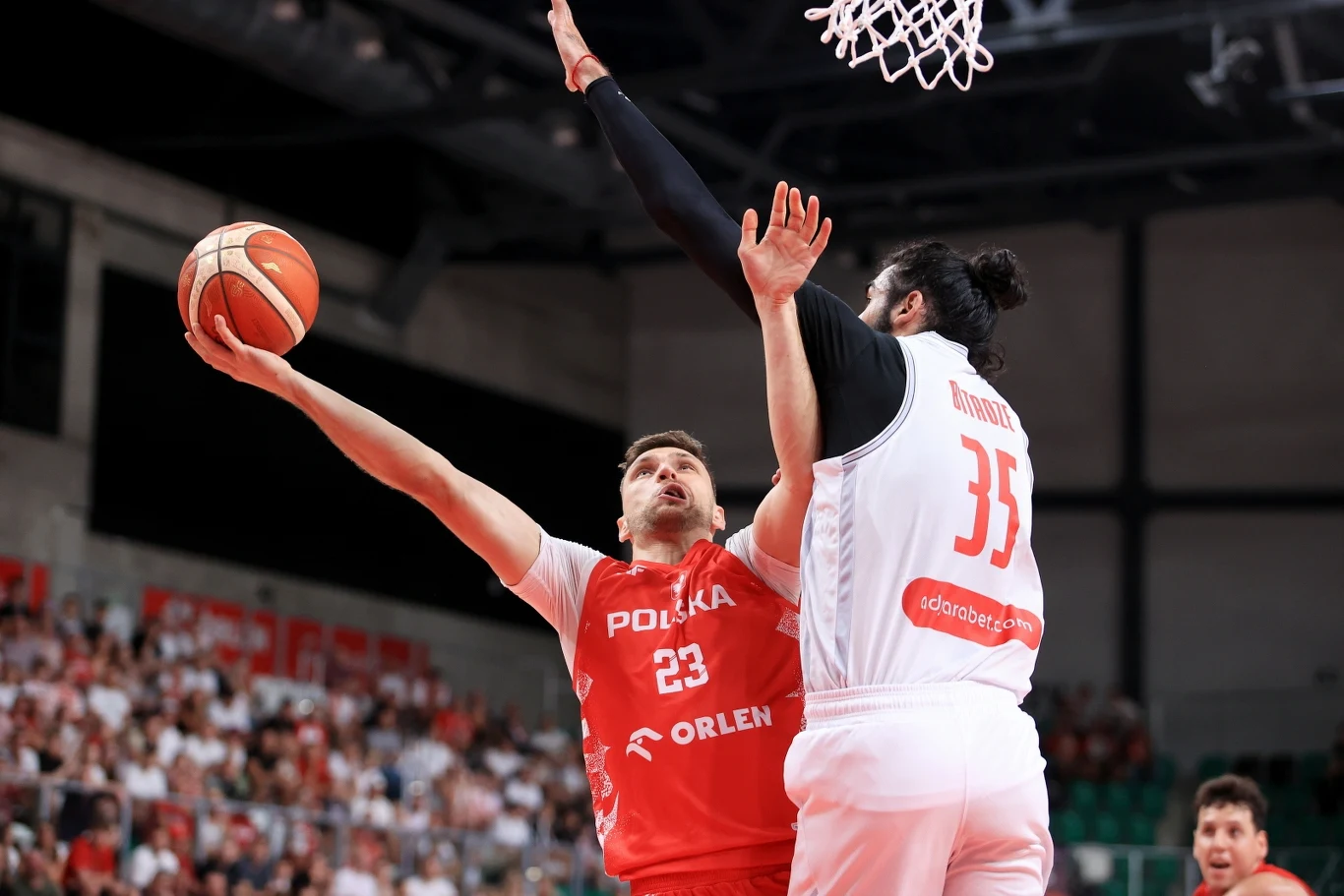 Michał Michalak okazał się superrezerwowym Zawodnik w czerwonym stroju z napisem 'Polska Orlen' wykonuje rzut do kosza, podczas gdy zawodnik drużyny przeciwników w białym stroju próbuje go zablokować, w tle widownia i kosz do koszykówki.