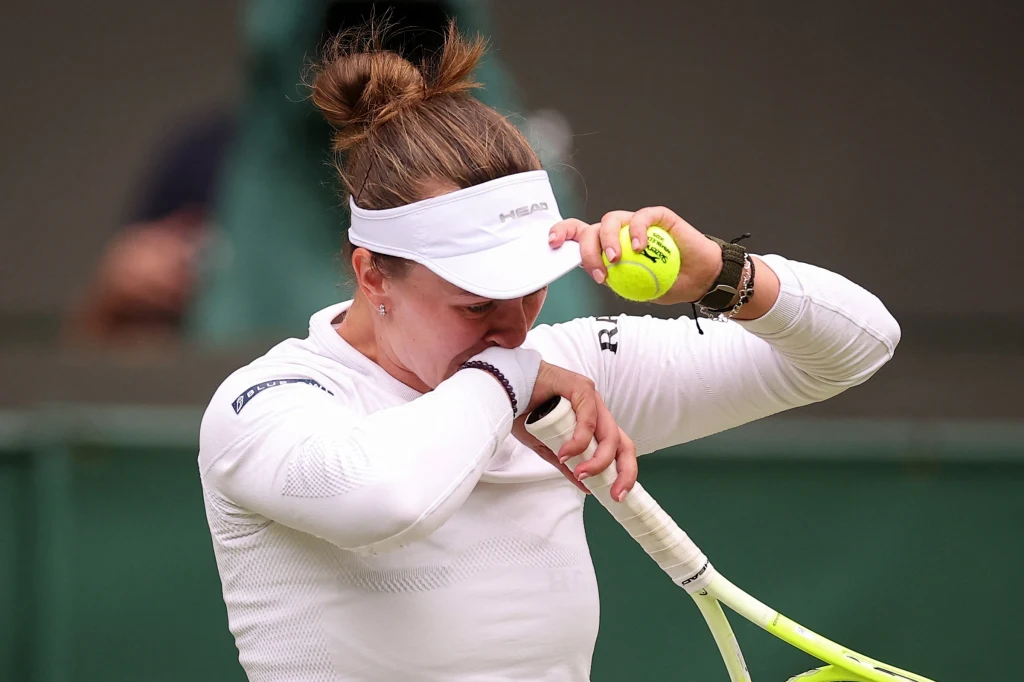 Barbora Krejcikova Tenisistka ubrana w biały sportowy strój i białą opaskę na głowie wyciera twarz ramieniem, trzymając w drugiej ręce rakietę tenisową i piłkę, autentyczny wyraz emocji podczas meczu.