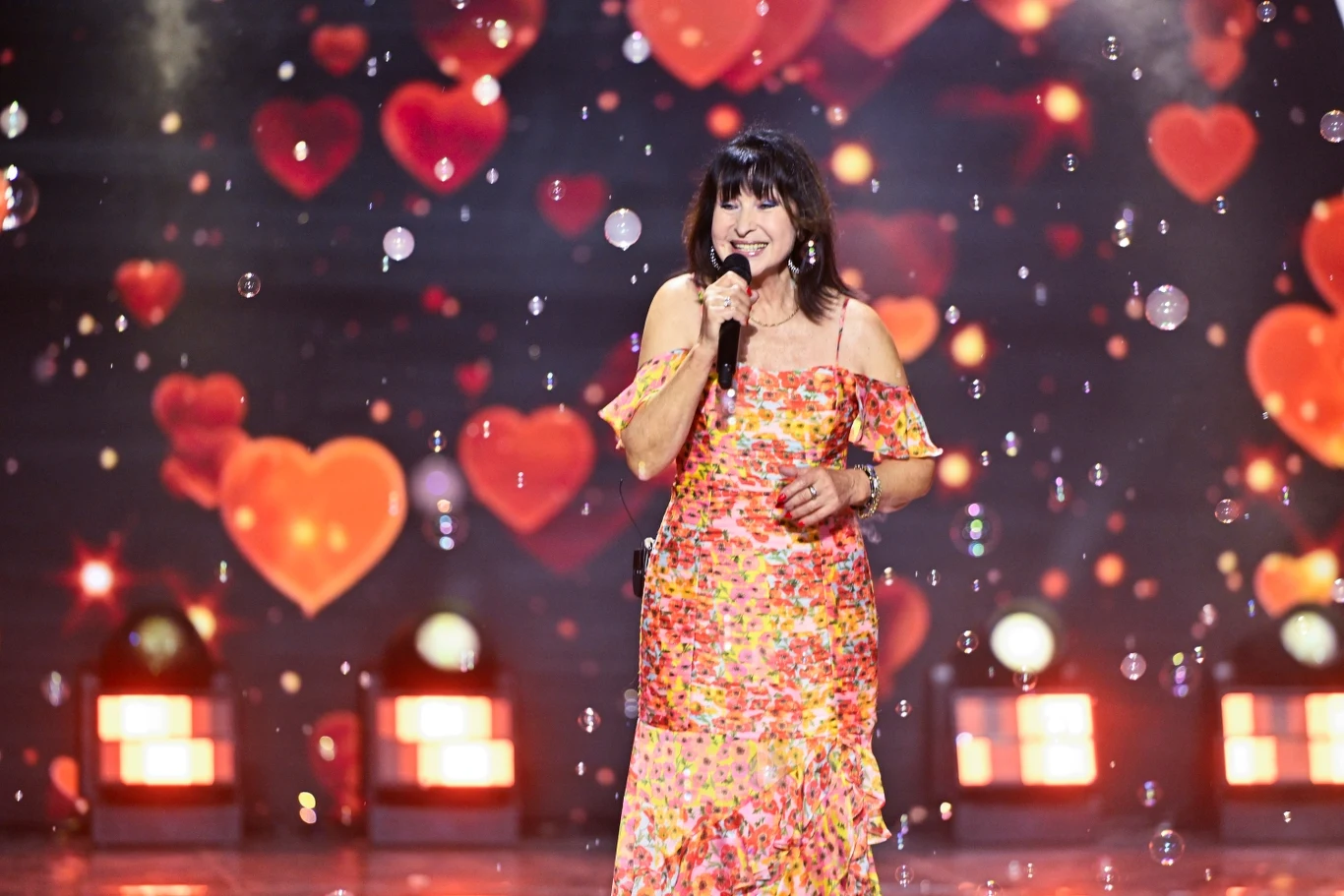 Tak wygląda obecnie Krystyna Giżowska Kobieta w kolorowej sukience śpiewa na scenie, trzymając mikrofon, w tle znajdują się liczne czerwone serca i światła sceniczne.