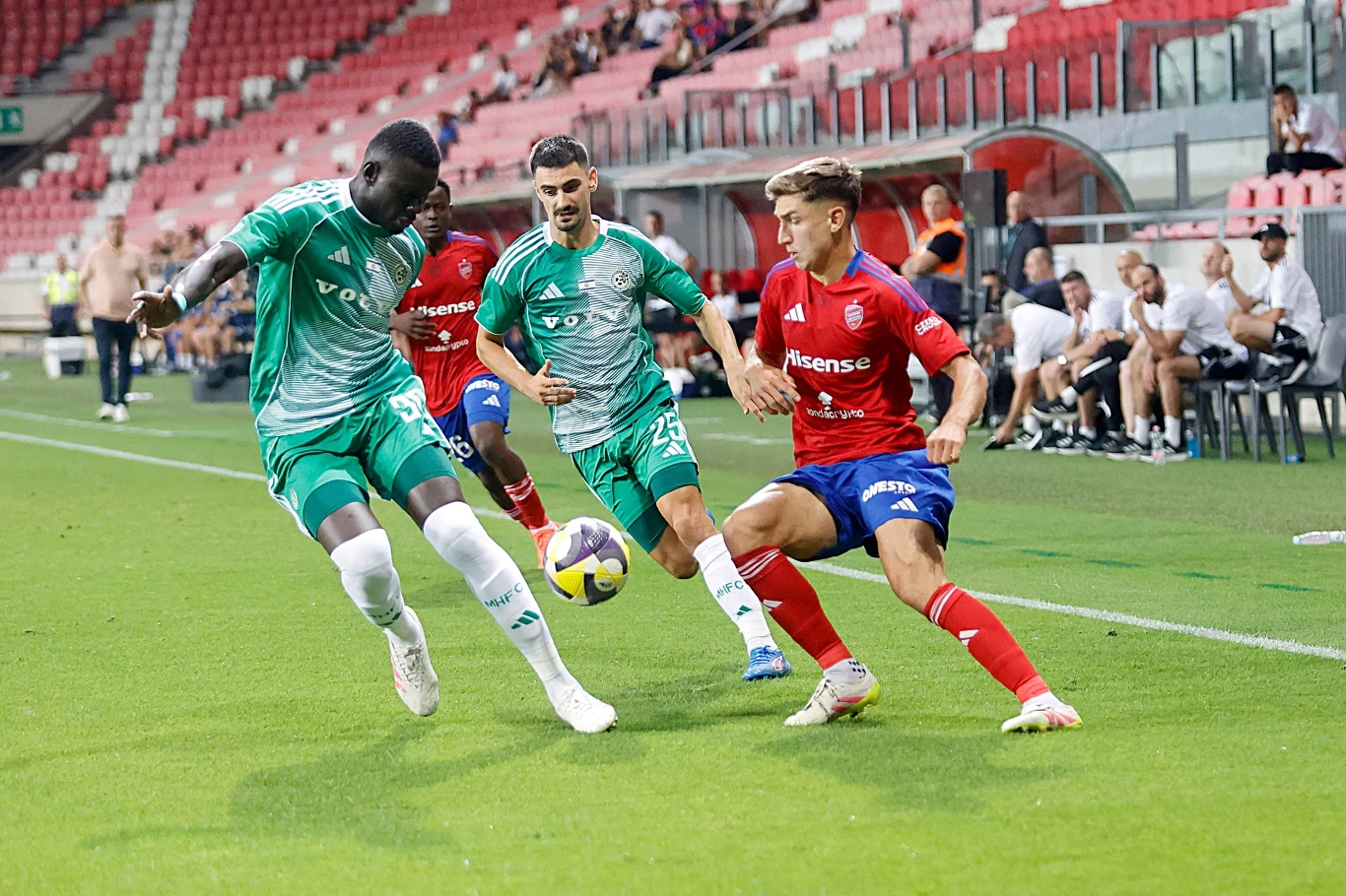 Maccabi Haifa - Raków Częstochowa Trzech piłkarzy rywalizuje o piłkę podczas meczu piłkarskiego na zielonym boisku, na trybunach widoczni są kibice i ławka rezerwowych.