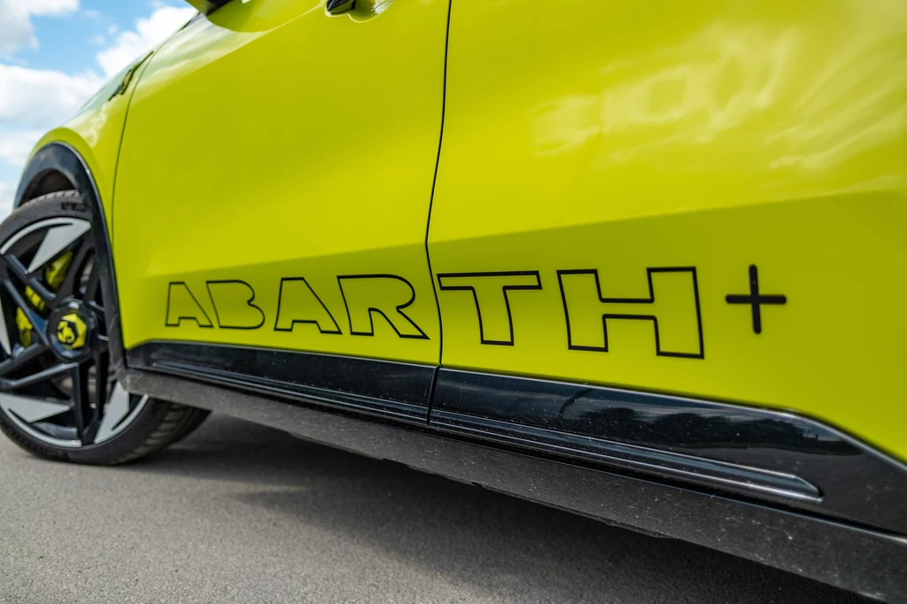 Abarth 600e Scorpionissima Bok żółtego samochodu sportowego z widocznym napisem ABARTH+, czarna felga i fragment linii drzwiowej, detal nad asfaltową nawierzchnią.