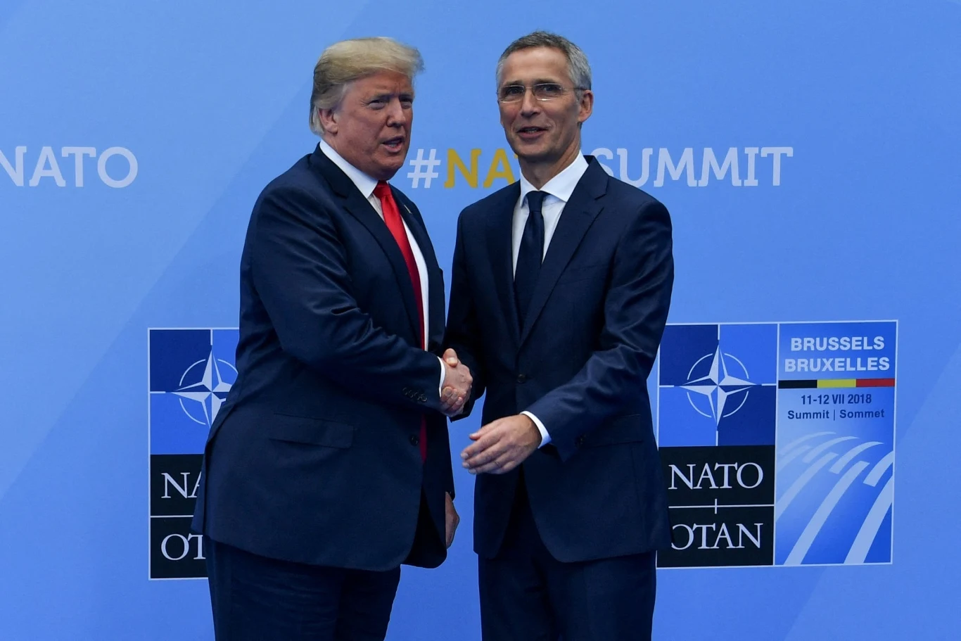 Donald Trump miał rozmawiać z Jensem Stoltenbergiem o Pokojowej Nagrodzie Nobla Dwóch mężczyzn w garniturach podaje sobie dłonie na tle niebieskiej ścianki z logo NATO oraz napisem NATO SUMMIT, z prawej strony znajduje się tablica informująca o szczycie NATO w Brukseli w 2018 roku.