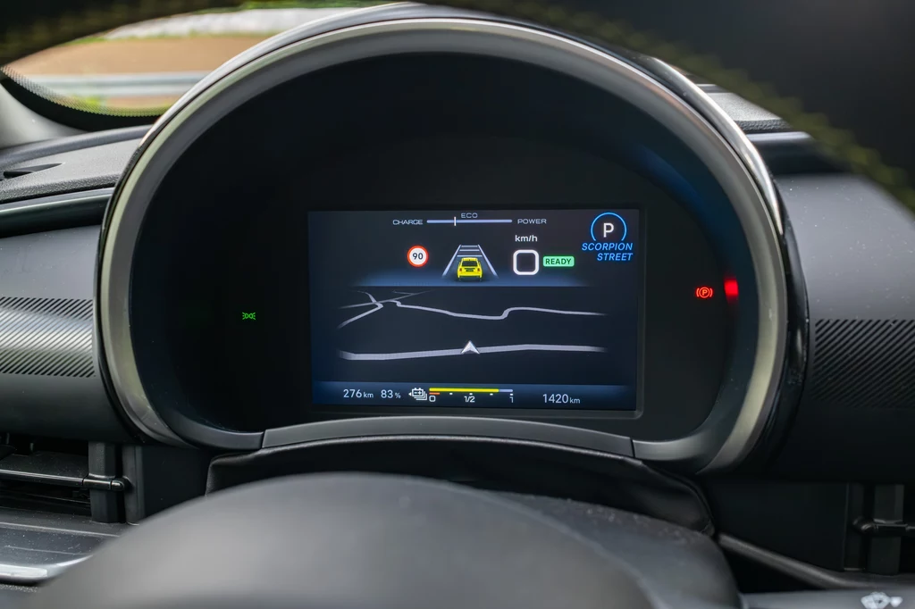 Abarth 600e Scorpionissima Cyfrowy kokpit samochodu z wyświetlaczem prezentującym informacje o systemie nawigacji, trybie jazdy, poziomie baterii i aktualnie aktywnych asystentach kierowcy.