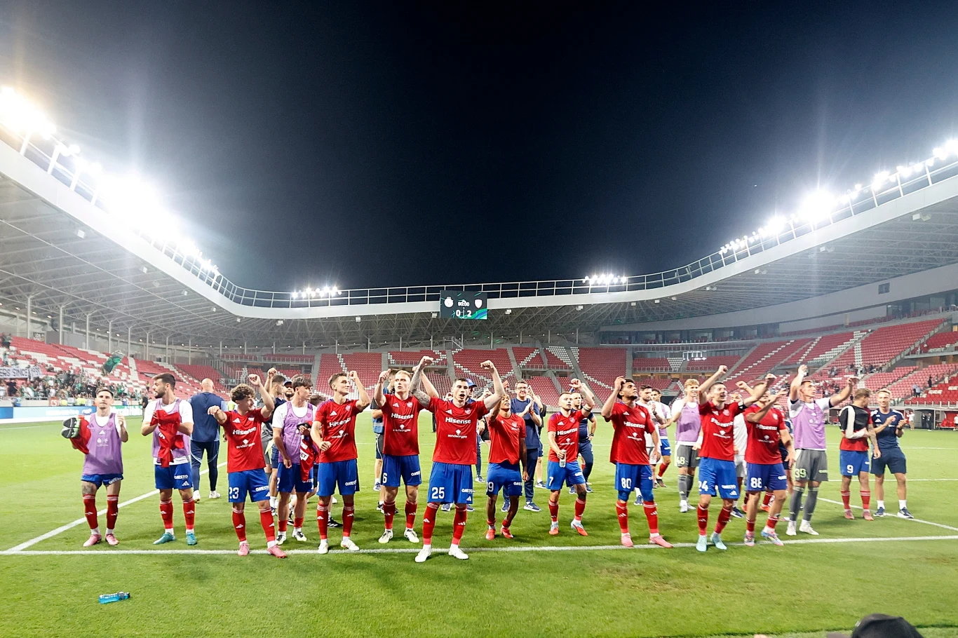 Drużyna piłkarska ubrana w czerwono-niebieskie stroje świętuje zwycięstwo na stadionie, podnosząc ręce do góry przed rozświetlonym trybunami, z tablicą wyników widoczną w tle.