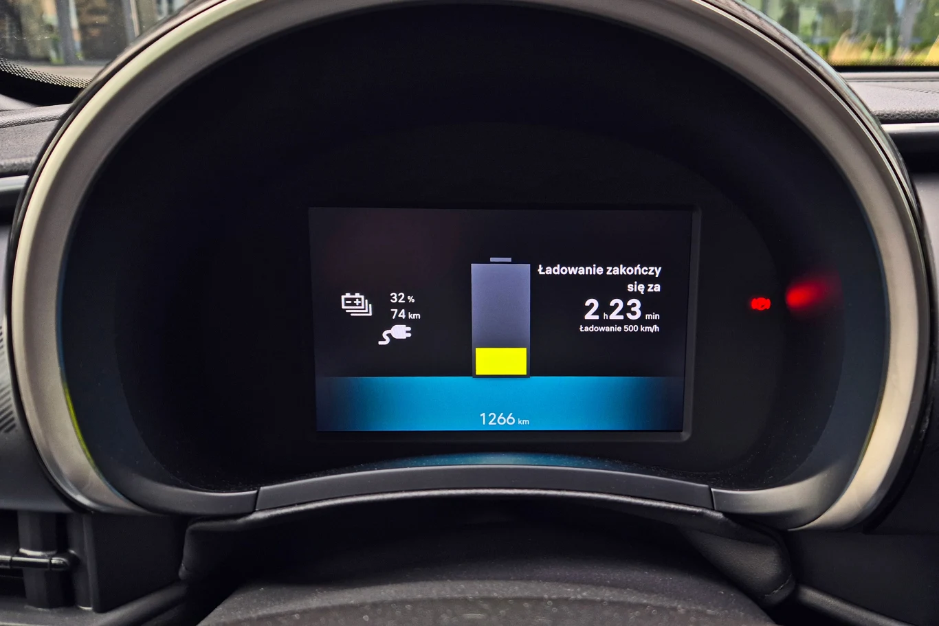 Kolorowy ekran deski rozdzielczej samochodu elektrycznego z czytelnym wskaźnikiem poziomu naładowania akumulatora na 32 procent, prognozowanym zasięgiem 74 km oraz czasem do zakończenia ładowania.