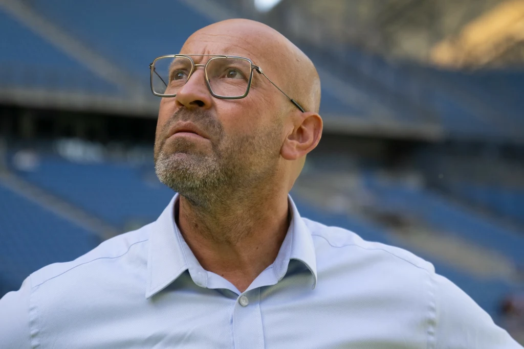 Łysy mężczyzna w okularach i jasnej koszuli patrzy w górę, stojąc na tle pustych, niebieskich trybun stadionu.