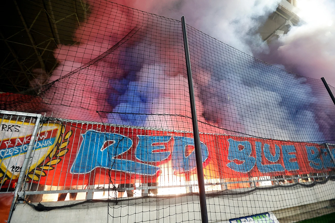 Kolorowe race dymne unoszą się nad trybuną kibiców, za siatką widoczny duży baner z napisem 'RED BLUE', na stadionie dominuje atmosfera sportowego dopingu.