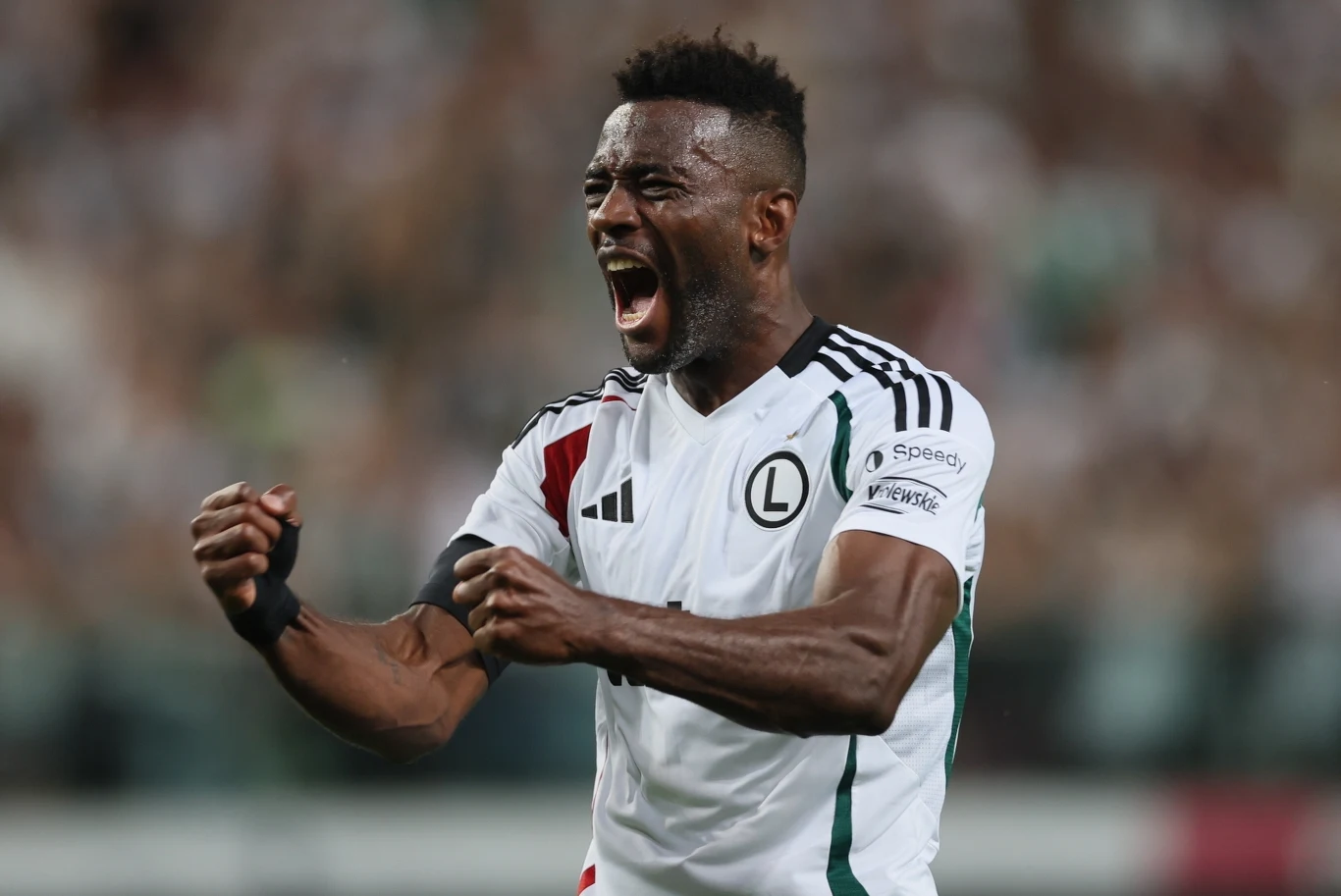 Legia jest wciąż w grze o Ligę Konferencji Piłkarz w białej koszulce z herbem Legii Warszawa świętuje zaciśniętymi pięściami i wyrażając ogromną radość, dynamiczne tło stadionu z kibicami jest rozmyte, skupienie na emocjach sportowca.