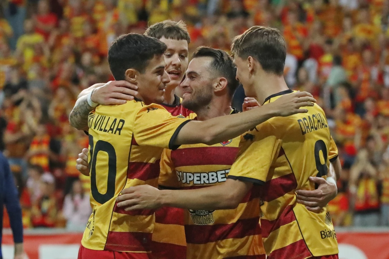 Jagiellonia awansowała do 4. rundy el. Ligi Konferencji Grupa piłkarzy w klubowych koszulkach świętuje wspólnie zdobycie gola na stadionie otoczonym przez kibiców w tle, zawodnicy obejmują się z uśmiechami na twarzach.