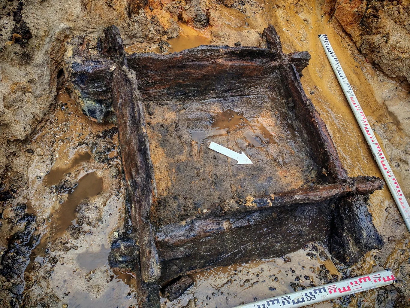Znalezisko na budowie drogi S8 Drewniana konstrukcja kwadratowa częściowo zanurzona w ziemi i błocie, otoczona wilgotnym podłożem, z białą strzałką wskazującą kierunek oraz miarą geodezyjną do pomiaru wykopów.