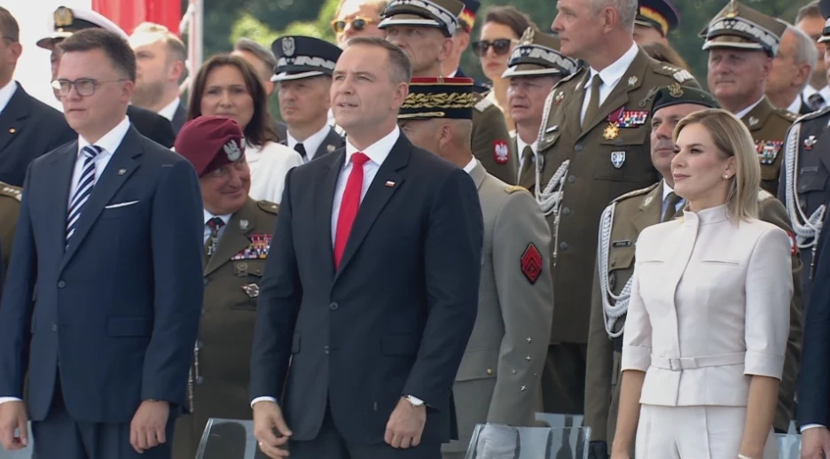 Grupa osób w oficjalnych strojach, wśród nich znajdują się wojskowi w mundurach oraz osoby cywilne, wszyscy stoją podczas oficjalnej uroczystości, część osób skupia uwagę na czymś niewidocznym poza kadrem.