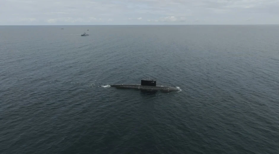 Okręt podwodny płynący po powierzchni spokojnego morza, w oddali widoczny inny okręt wojskowy na horyzoncie pod zachmurzonym niebem.