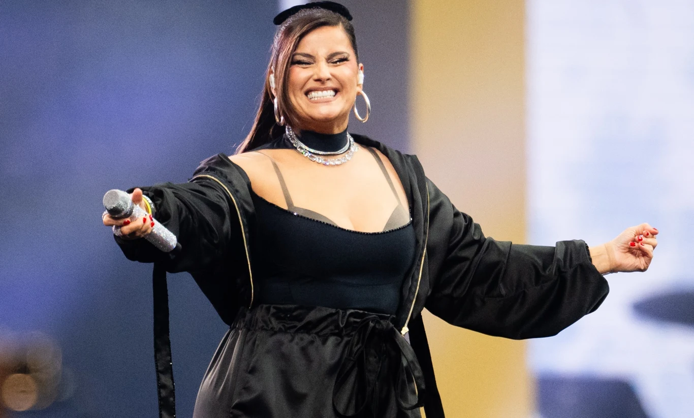 Nelly Furtado była jedną z gwiazd Bittersweet Festival Kobieta na scenie w czarnym stroju i płaszczu, z szerokim uśmiechem trzyma mikrofon, za nią rozmyte tło z jasnym fragmentem.