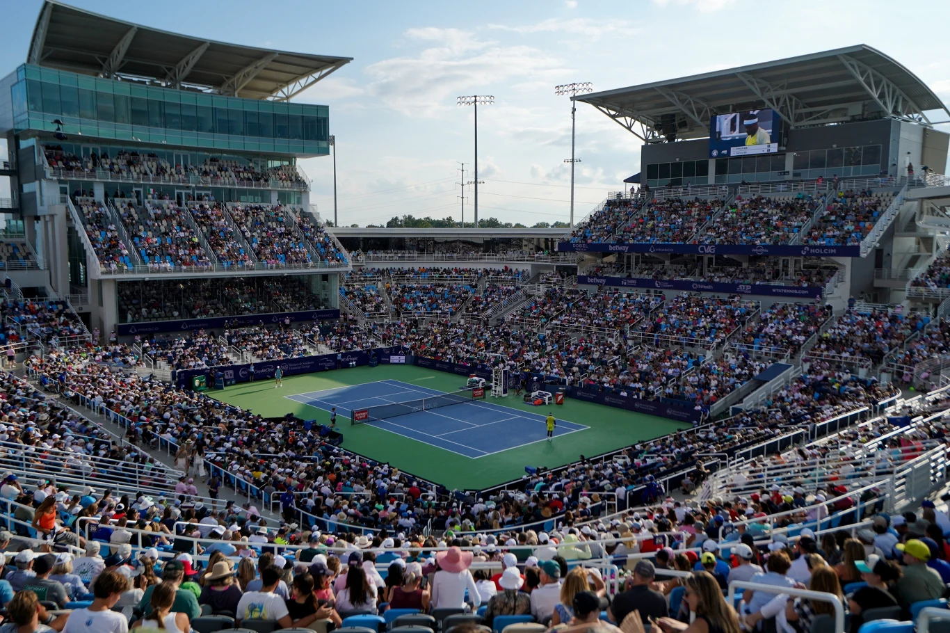 Tenis Kort tenisowy na otwartym stadionie wypełnionym publicznością, na środku rozgrywany jest mecz tenisowy, a widzowie zajmują miejsca na trybunach wokół boiska.