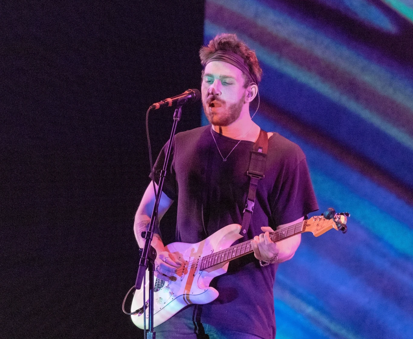 Two Feet Młody mężczyzna śpiewający do mikrofonu i grający na gitarze elektrycznej na scenie, dynamiczne kolorowe światło w tle dodaje energii występowi.